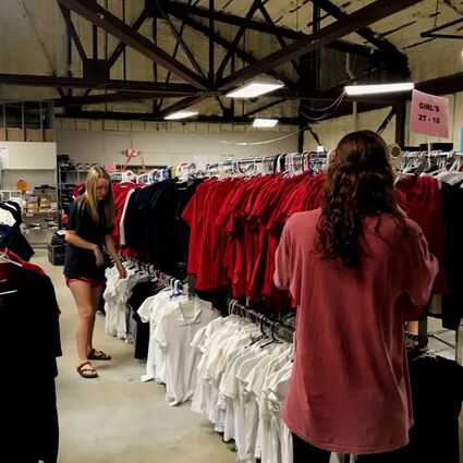 Two young girls are organizing the uniform polos that hang on long clothing racks.